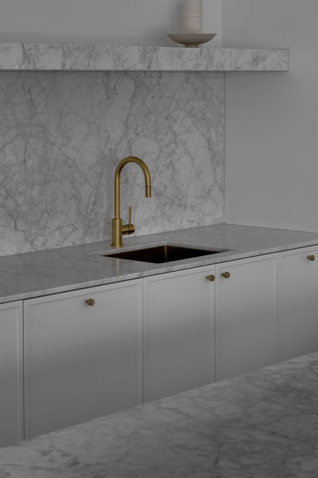 Close-up view of a refined kitchen with pale grey veined marble benchtops and full-height marble splashback, white shaker-style cabinetry with small brass knobs, and a curved brass gooseneck mixer tap above an undermount sink. A slim marble shelf with a simple decorative vessel runs along the upper wall. The overall palette is soft grey, white, and brass with a restrained, classic feel.
