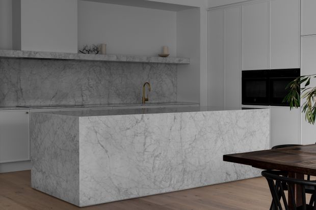 A refined contemporary kitchen featuring a large full-height marble-look island with waterfall ends, matching marble splashback and rear benchtop, white panelled cabinetry, integrated black appliances, and a brass-toned tap. Warm timber flooring and a dark timber dining table add contrast. The space is light, clean, and softly minimalist.
