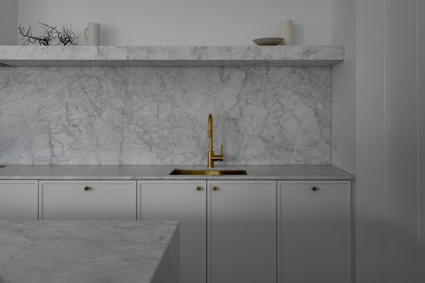 A refined kitchen joinery detail in a muted grey-white palette, featuring extensive veined marble on the benchtops and backsplash, pale grey shaker-style cabinetry with small dark knobs, and a brass gooseneck mixer tap. A marble shelf with minimal ceramic decor runs above the splashback, while a foreground island is partially visible.