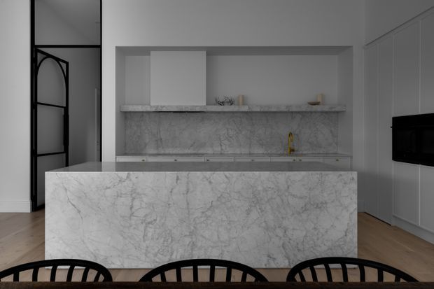 A refined contemporary kitchen viewed from the dining area, featuring a large marble-clad island, matching marble splashback and benchtop, white integrated cabinetry, a concealed rangehood, and a small gold-toned tap. Pale timber flooring and black-framed glazed internal doors add contrast.