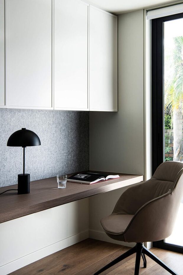 A compact, refined home office nook with a floating dark timber desk integrated beneath white overhead cabinetry. The workspace includes a black dome desk lamp, open book, and glass, with a sculptural taupe upholstered swivel chair beside full-height black-framed glazing. A speckled grey backsplash or wall panel sits behind the desk, and warm timber flooring continues below.