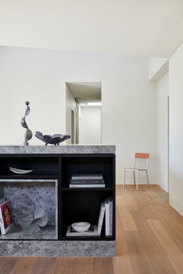 A bright, minimalist interior hallway or living-area threshold featuring a dramatic black built-in shelving unit with grey veined stone cladding and open display compartments. Light oak timber flooring, white walls and ceiling, and a simple orange-and-metal chair create a restrained contemporary composition. Sculptural objects and books are arranged on the shelving, with a corridor visible beyond.