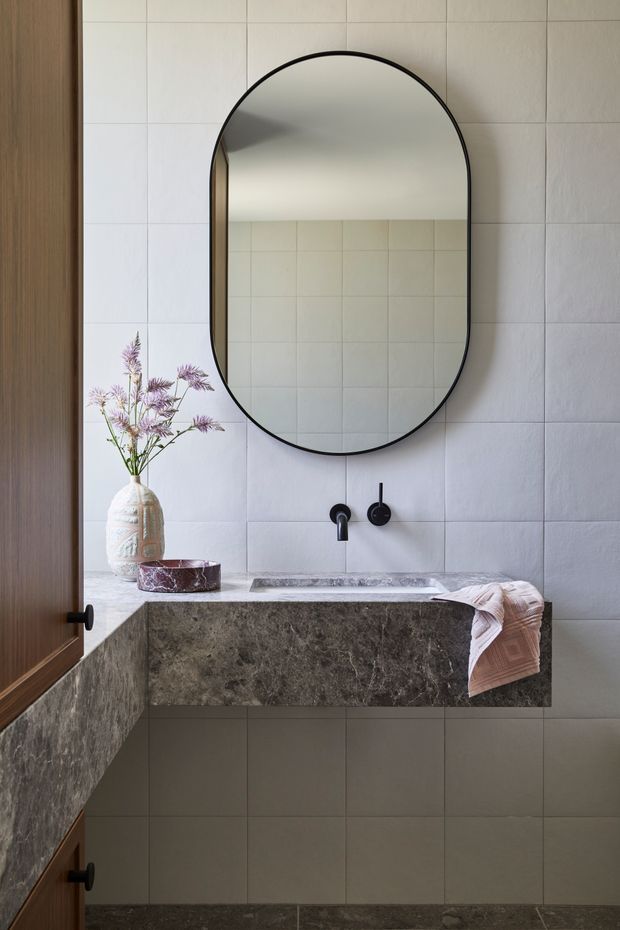 A refined bathroom vanity with a large rounded-arch black-framed mirror, pale square wall tiles, a dark stone floating benchtop and integrated basin, and matte black wall-mounted tapware. Timber cabinetry frames the left side, while a ceramic vase and folded towel add subtle decorative accents.
