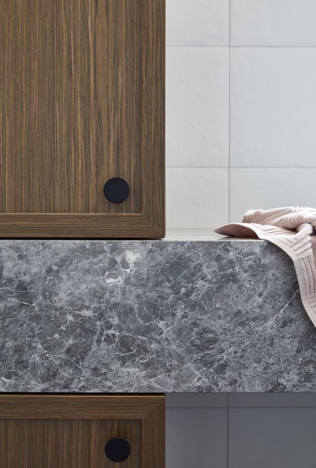 Close-up detail of a refined bathroom vanity or built-in cabinet, featuring dark walnut-toned timber cabinetry with black round knobs, a dramatic grey veined stone benchtop, and pale grey tiled wall surfaces. A folded blush-beige towel rests on the benchtop, adding a soft accent.