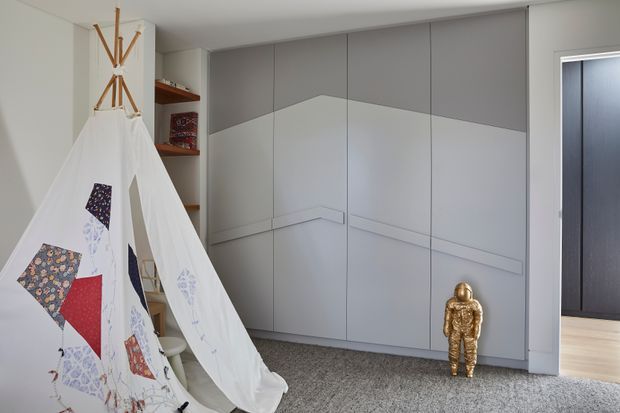 A bright, modern children's bedroom featuring a large white fabric teepee with colourful patchwork panels, full-height pale grey built-in wardrobes with geometric decorative panelling, open shelving, grey carpet, and a doorway leading to a darker adjacent space. A small gold decorative figure stands in front of the wardrobes.