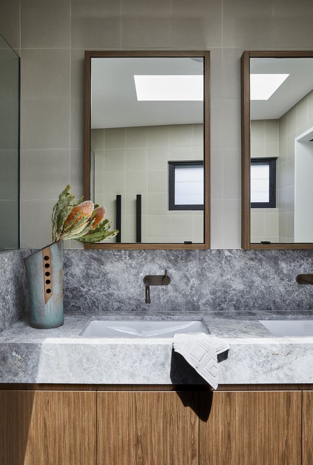 Contemporary bathroom vanity with a double basin, grey veined stone benchtop and splashback, timber cabinetry, dark-framed mirrors, wall-mounted black taps, and pale tiled walls. A skylight and reflected window bring natural light into the space.