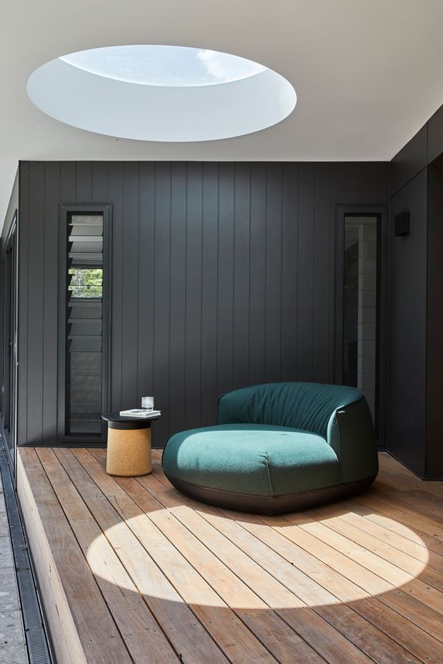 A compact, refined lounge area with a deep teal upholstered lounge chair and small cylindrical side table, set against dark vertical wall panelling. Warm timber decking covers the floor, while a large circular skylight creates a dramatic pool of sunlight. Narrow louvred windows and dark built-in wall surfaces add a modern architectural character.