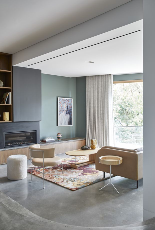 Refined contemporary living room with a built-in fireplace and media wall, timber cabinetry, muted green walls, polished concrete flooring, and large windows dressed with full-height curtains. A tan sofa, lounge chair, round coffee table, side table, and colourful patterned rug create a warm, curated seating area. The room features a clean-lined recessed ceiling detail and abundant natural light.