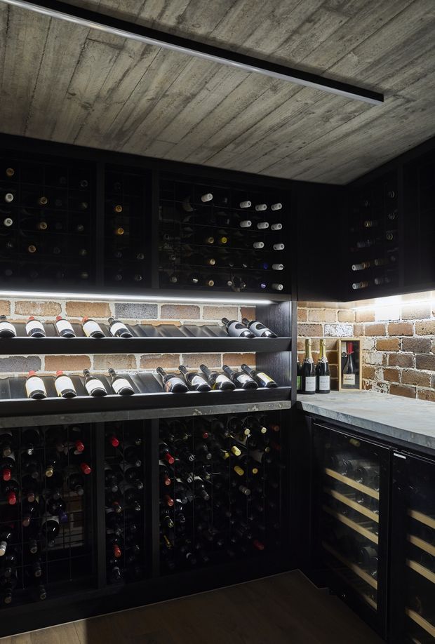 A compact, dark wine cellar with extensive black built-in bottle storage, horizontal wine display shelves, under-counter wine refrigerators, a grey stone benchtop, exposed textured brick walls, and a raw exposed concrete ceiling. Linear integrated lighting highlights the bottles and shelving.