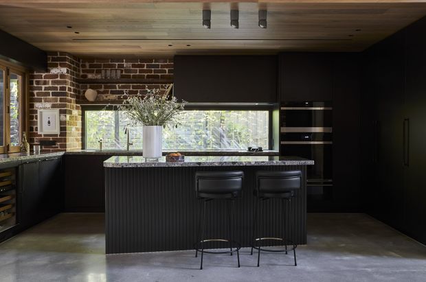 A dark, sophisticated contemporary kitchen with black cabinetry, a large central island with two bar stools, integrated ovens, and a long horizontal window. Exposed red brick forms a feature wall, while the ceiling is clad in warm timber boards and the floor is polished concrete. Natural greenery is visible through the glazing.