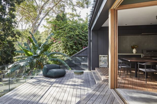 A contemporary elevated timber deck adjoining an open-plan dining area through large timber-framed sliding doors. The deck has grey timber decking, frameless glass balustrades, a rounded dark lounge chair and small side table, with dense leafy landscaping beyond. Inside, a timber dining table and chairs sit beneath a dark ceiling and beside dark built-in cabinetry.