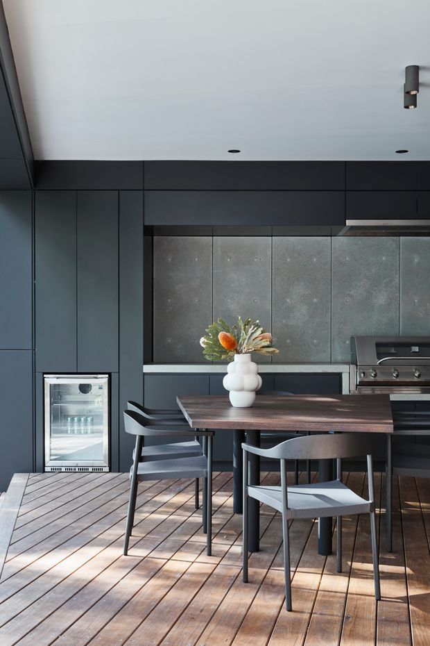A dark, sophisticated contemporary kitchen and dining area with full-height charcoal cabinetry, a concrete-look tiled splashback, integrated outdoor-style grill, under-counter beverage fridge, and a dark timber dining table surrounded by modern black chairs. The room opens onto a timber deck-like floor and is lit by recessed ceiling lights and natural daylight.