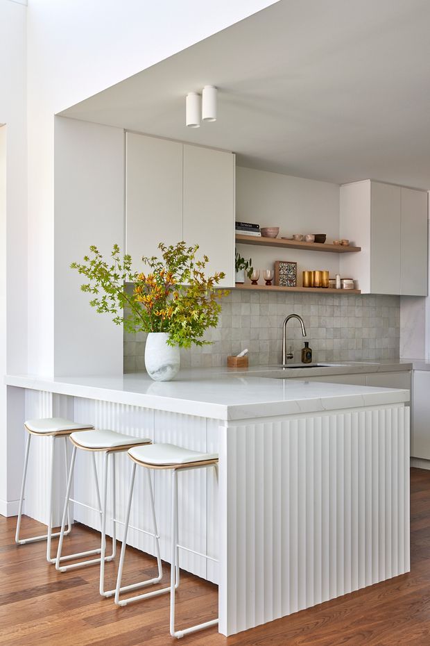 Bright, refined contemporary kitchen featuring a large white island with a vertically fluted panelled base and waterfall-style benchtop, three slim bar stools, pale grey square-tile splashback, integrated white cabinetry, open timber shelving, and brushed metal tapware. Warm timber flooring and carefully styled ceramics and greenery add softness to the predominantly white scheme.