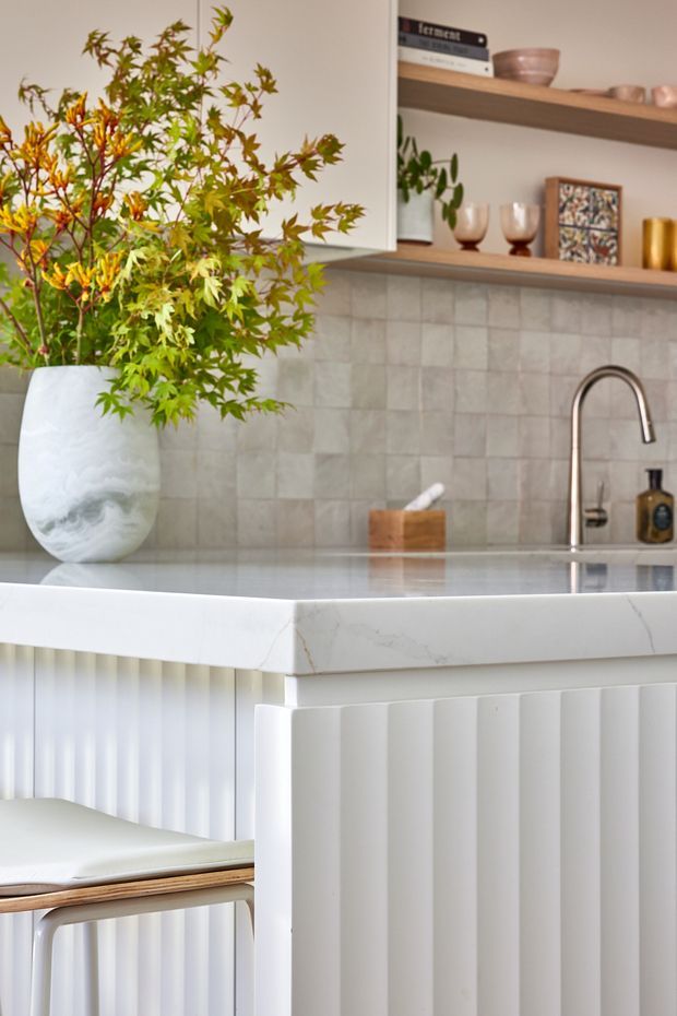 A bright, refined kitchen detail featuring a white marble-look island benchtop with a vertically fluted white base, a pale square-tile splashback, open timber shelving, and a brushed metal gooseneck tap. Decorative ceramics, books, and a large leafy arrangement add warmth and texture.