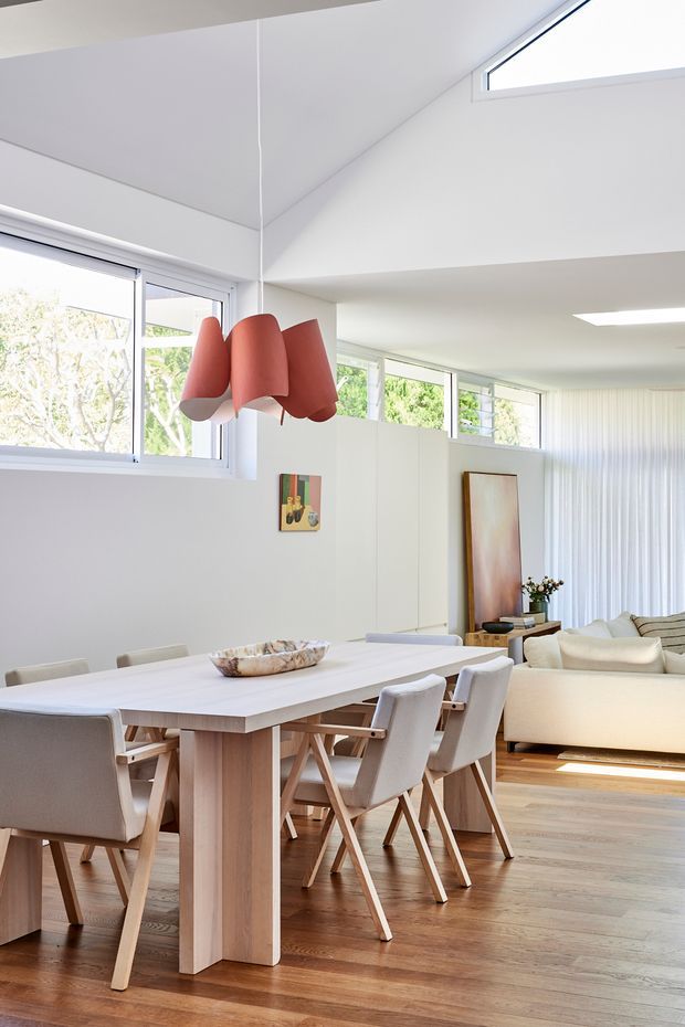 Bright open-plan dining and living area with a large pale timber dining table, upholstered dining chairs, white modular sofa, timber flooring, extensive windows, and a sculptural coral pendant light. The room has a high angled ceiling and a clean, contemporary coastal feel.