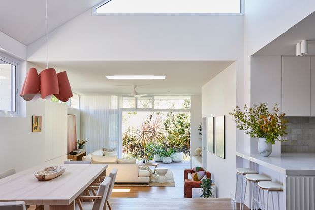 Bright open-plan contemporary interior combining a dining area, living room and kitchen. The space features white walls and cabinetry, pale timber flooring, a large skylight, clerestory-style glazing, expansive rear windows, a sculptural red pendant light, light upholstered furniture, and a compact kitchen island with stools. Indoor plants and garden views add a fresh, airy atmosphere.