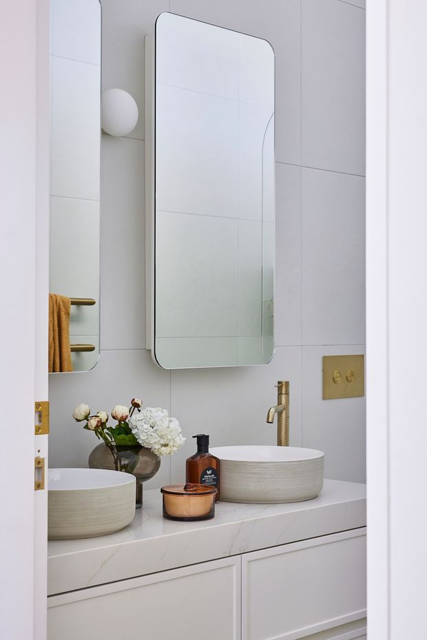 A refined contemporary bathroom viewed through a partially open door, featuring a double vanity with two round vessel basins on a pale stone benchtop, integrated white cabinetry, large-format light wall tiles, rounded rectangular mirrors, and brushed brass tapware and hardware. Soft floral arrangements and warm amber accessories add subtle contrast to the light neutral scheme.