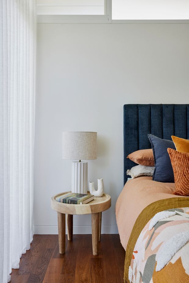 Bright, softly styled bedroom corner featuring a tall vertically channelled navy upholstered headboard, layered rust, blue, ochre and blush bedding, a round timber bedside table with a fabric-shaded lamp and small ceramic vessels, dark timber flooring, pale walls, and full-height sheer curtains beside a high window.