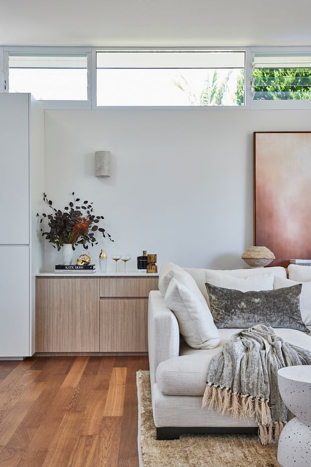 Bright, refined living room with white walls, high horizontal windows, warm timber flooring, and a light upholstered sofa layered with textured cushions and a throw. A low timber-grain sideboard provides storage beneath minimalist decor and dried botanicals, while a large blush-toned artwork adds a soft feature accent. The room has an airy, contemporary residential interior with abundant natural light.