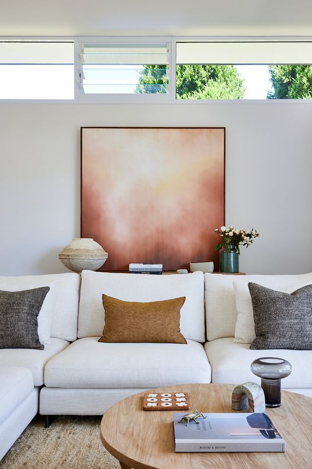 Bright, refined living room with a large off-white sectional sofa, textured neutral cushions, a rounded timber coffee table, and a warm abstract artwork above a slim console. High horizontal windows with white frames and blinds bring in daylight, while ceramic vessels, a glass vase, books, and a small decorative object add understated styling.