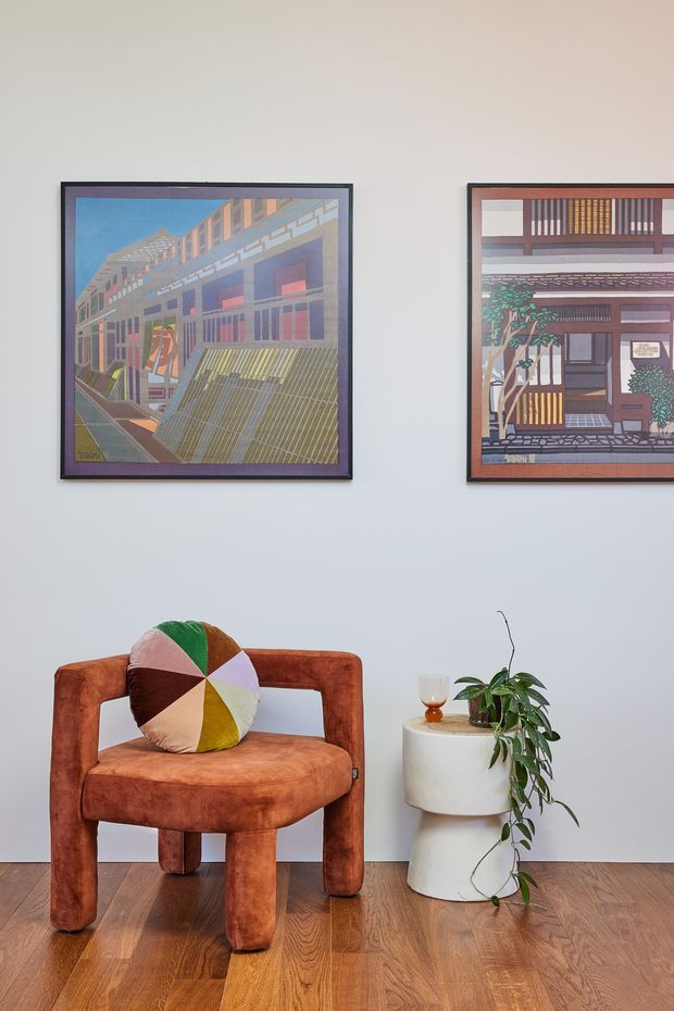 A styled interior vignette with a rust-orange upholstered lounge chair, multicoloured geometric cushion, and a small white sculptural side table holding a potted trailing plant and glass. Two framed architectural artworks hang on a pale wall above warm timber flooring.