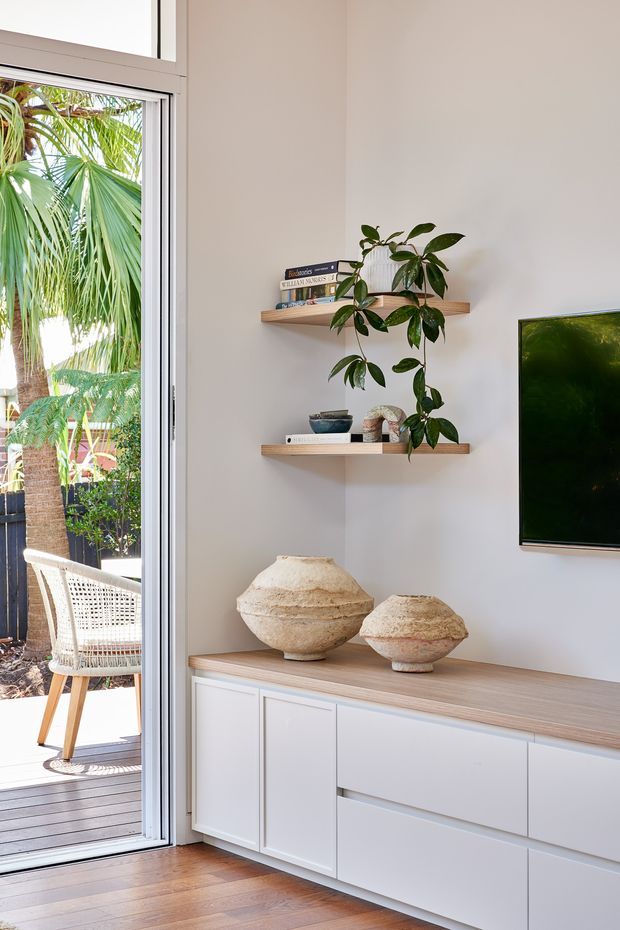 Bright contemporary living room corner with a white built-in media console, pale timber benchtop, floating timber display shelves, decorative ceramic vessels, indoor plants, and a wall-mounted television. A large glazed sliding door opens to a timber deck and lush tropical planting outside.