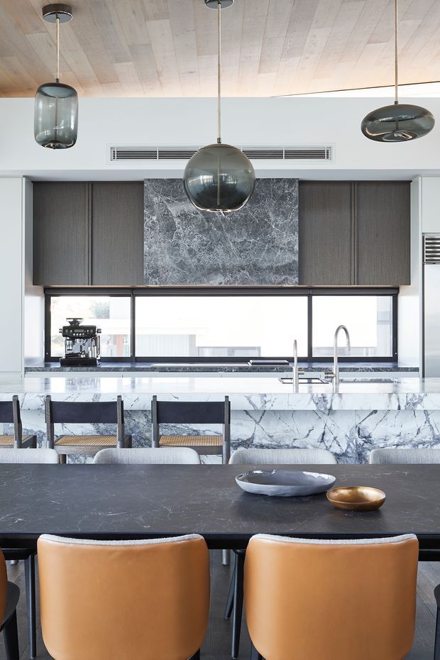 A sophisticated open-plan kitchen and dining area featuring a large marble-clad island with integrated seating, dark timber cabinetry, a dramatic grey marble backsplash, and expansive black-framed windows. The ceiling is lined with pale timber boards and illuminated by sculptural smoked-glass pendant lights. In the foreground, a dark dining table is paired with warm tan leather dining chairs.