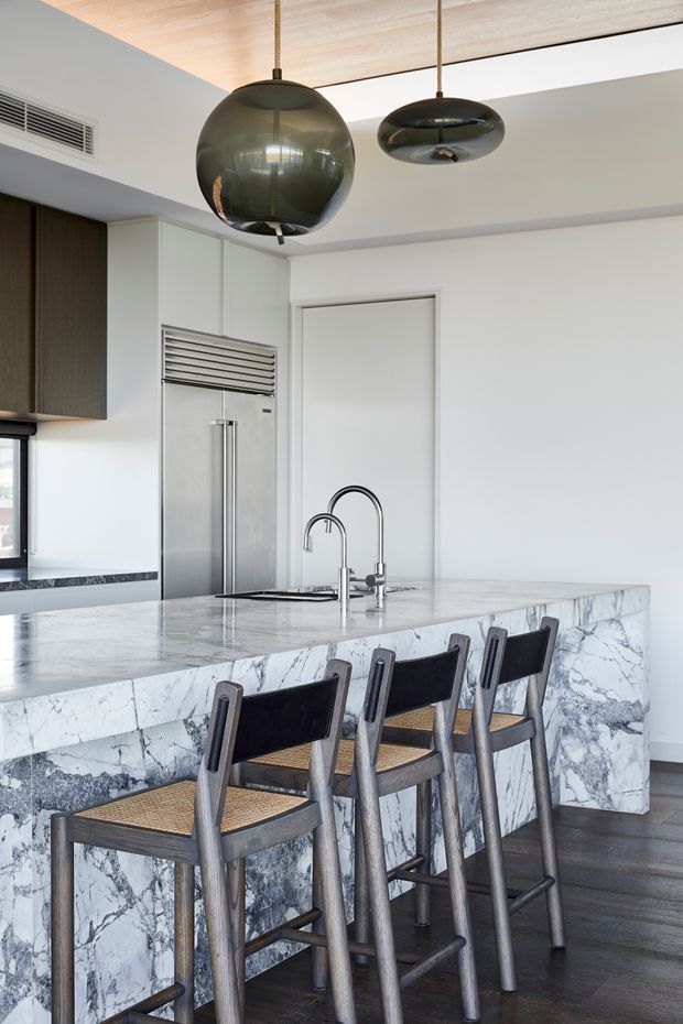 A bright contemporary kitchen centered on a large marble waterfall island with an integrated sink and twin curved faucets. Four timber-framed bar stools with dark backs and woven seats line the island. White cabinetry, a stainless-steel refrigerator, dark timber flooring, and two sculptural pendant lights complete the refined interior.
