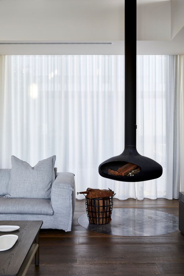 A refined living room with a pale grey sofa, sheer full-height curtains, dark timber flooring, and a dramatic suspended black fireplace with a visible log basket beneath it. The space is softly lit and minimally furnished, with a neutral palette and contemporary detailing.