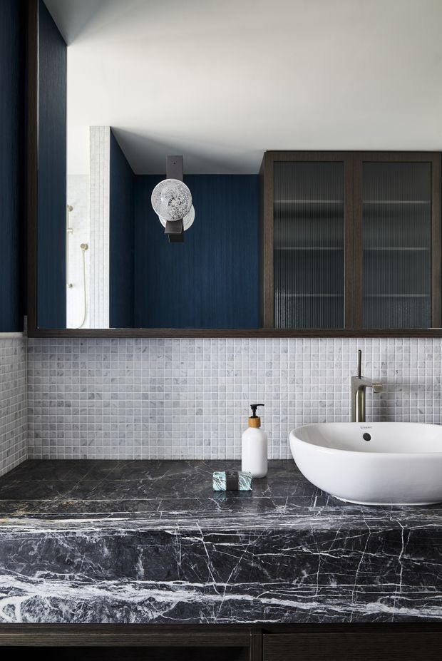 A refined contemporary bathroom vanity viewed straight on. The room features a white vessel basin set on a dramatic dark veined stone benchtop, a small pale grey mosaic-tile splashback, dark timber vanity cabinetry, a brushed metallic tap, a large mirror, and integrated dark-framed ribbed-glass storage cabinets. A spherical wall light and simple toiletries add subtle detail.