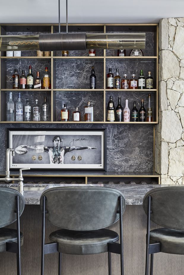 A sophisticated home bar with dark marble-look wall panels, brass-trimmed open shelving displaying liquor bottles and glassware, a framed monochrome artwork, and a stone-clad column. A marble or stone-topped bar counter is paired with dark leather bar stools and a large cylindrical pendant light. The palette is dark, refined, and masculine with metallic accents.
