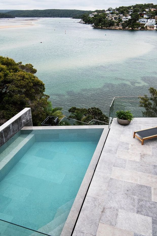 Elevated coastal pool terrace overlooking a calm turquoise bay, with a rectangular swimming pool, pale stone paving, frameless glass balustrades, a sun lounger and potted greenery. Houses are visible on the wooded shoreline across the water.