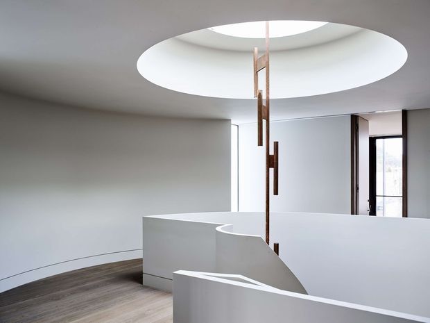 A minimalist interior landing or stairway with curved white balustrades and walls, dark timber flooring, and a dramatic circular skylight above. A vertical sculptural pendant or timber feature hangs through the skylight opening, while narrow dark-framed glazing is visible at the far end.
