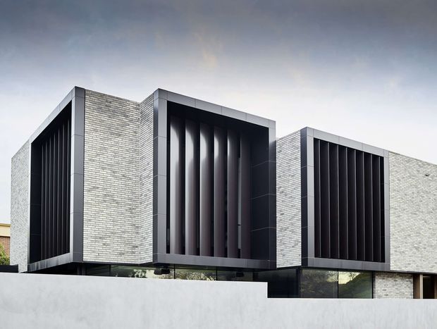 Contemporary multi-level residential facade with pale grey brick walls, strongly projecting dark-framed window bays, and deep vertical black louvres or fins screening the glazing. The composition is highly geometric, with flat rooflines, crisp rectilinear volumes, and limited visible landscaping behind a light boundary wall.