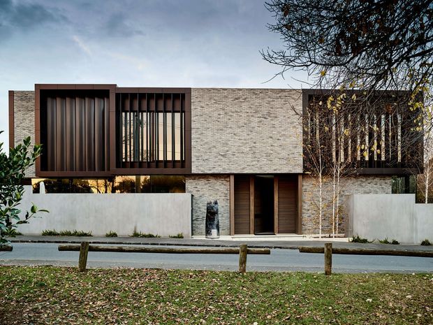 Front exterior view of a contemporary two-storey residence with a long, low-profile form. The facade is predominantly pale textured brick, complemented by dark timber or metal-framed recessed window screens with vertical slats, deep reveals, and warm-toned glazing. A sheltered central entry is set behind a low concrete boundary wall, with landscaped planting and a grass verge in front.
