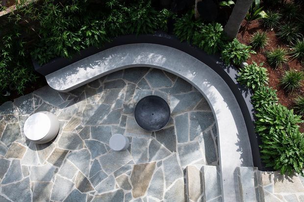 Aerial view of a landscaped curved outdoor terrace or courtyard with irregular natural stone paving, a sweeping smooth concrete bench or retaining edge, rounded outdoor stools or tables, steps, and dense tropical planting.