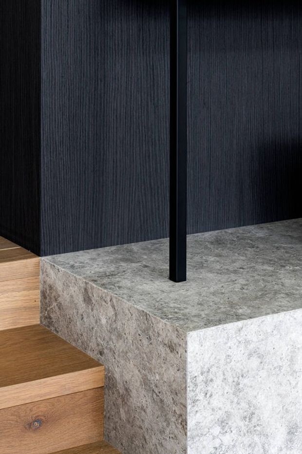 Close-up architectural detail showing a stair landing or raised platform clad in textured grey natural stone, adjacent to warm timber stairs and dark vertically textured wall panelling. A slim black structural or balustrade post rises from the stone surface, creating a refined contemporary interior composition.