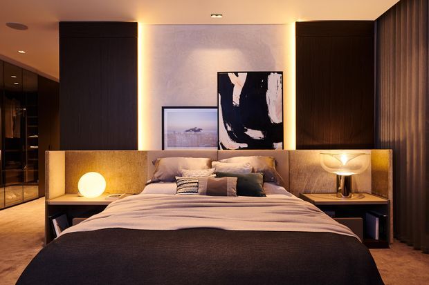 A warm, moody contemporary bedroom with a centrally positioned upholstered bed, layered neutral bedding, dark timber built-in cabinetry, integrated bedside surfaces, and softly illuminated wall panelling. Abstract artwork and ambient bedside lighting create a refined hotel-like atmosphere, with dark curtains and a glimpse of adjacent built-in storage at the left.