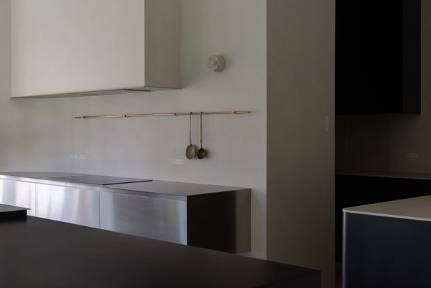 Minimalist contemporary kitchen with flat-panel stainless-steel cabinetry, a dark island or benchtop in the foreground, pale walls, and a sleek integrated rangehood. A slim brass rail with hanging utensils provides a small accent, while dark openings and cabinetry add contrast.