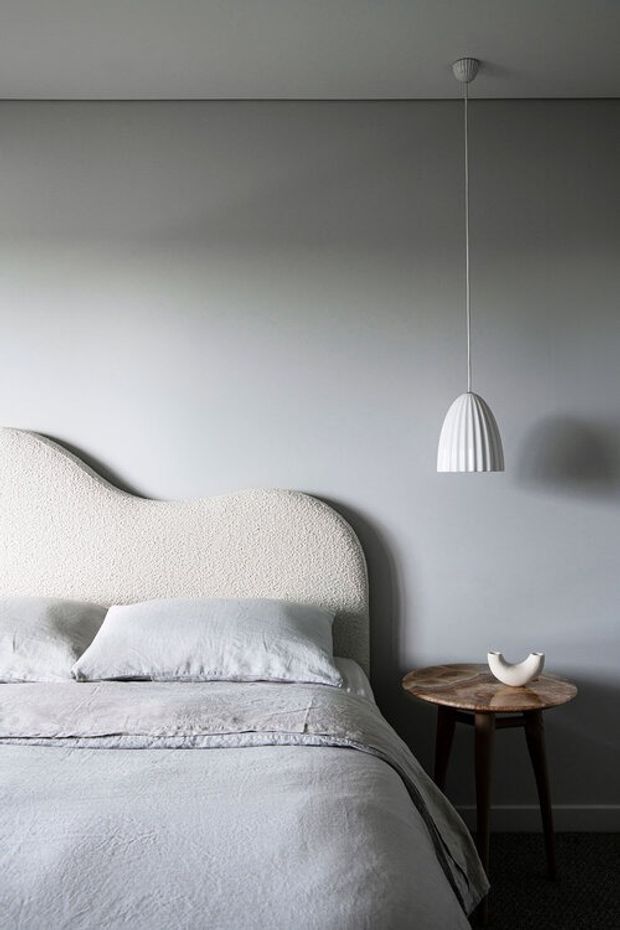 A softly lit, minimalist bedroom with a sculptural cream upholstered headboard, pale grey bedding, and a small dark wood side table topped with a simple white ceramic object. A ribbed white pendant light hangs beside the bed against a smooth grey wall, creating a calm and refined composition.