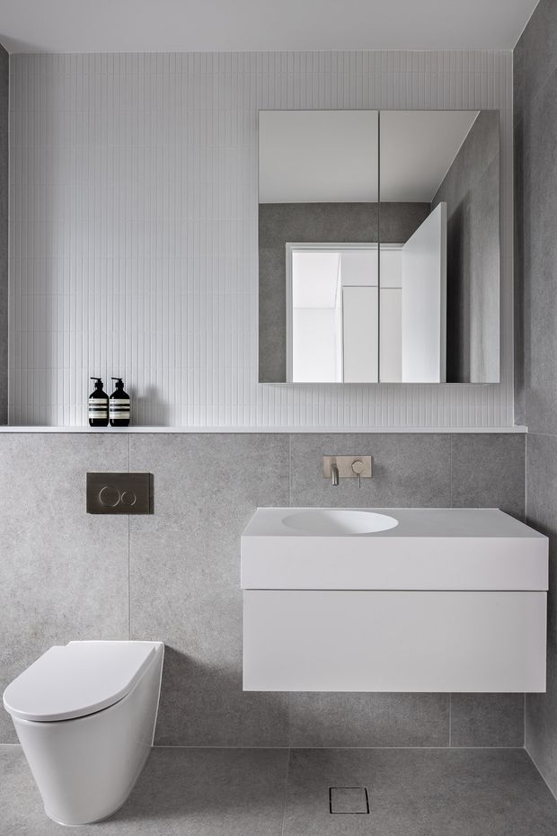 A compact contemporary bathroom in a restrained grey-and-white palette. Large grey wall and floor tiles are paired with a vertically fluted white tile feature wall, a wall-hung white vanity with integrated basin, mirrored cabinet, concealed cistern toilet, and minimalist brushed metal fittings. The room has a clean, refined, highly functional appearance.