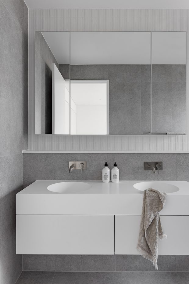 A contemporary minimalist bathroom featuring a floating double vanity with integrated white basins, wall-mounted chrome tapware, grey large-format tiled walls and floor, and a wide mirrored cabinet above a narrow vertical-ribbed tile or panel backsplash. Neutral grey and white finishes create a clean, refined appearance.