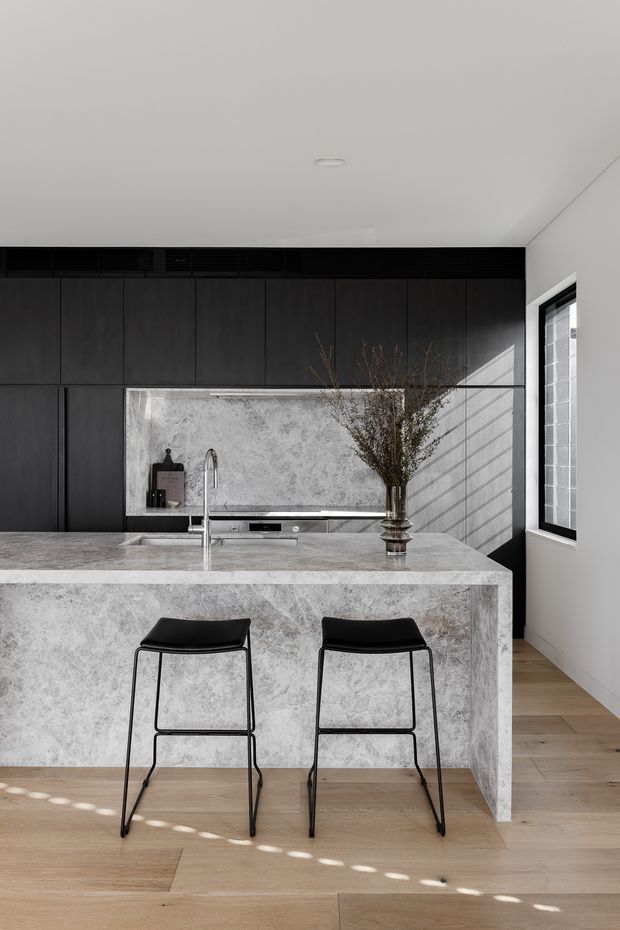 A refined contemporary kitchen with a large pale stone island and integrated waterfall edge, black bar stools, dark full-height cabinetry, and a matching stone splashback. A stainless-steel sink and tap sit within the island, while timber flooring, a high white ceiling, and a tall glazed window add warmth and natural light. A vase of branches provides a restrained decorative accent.