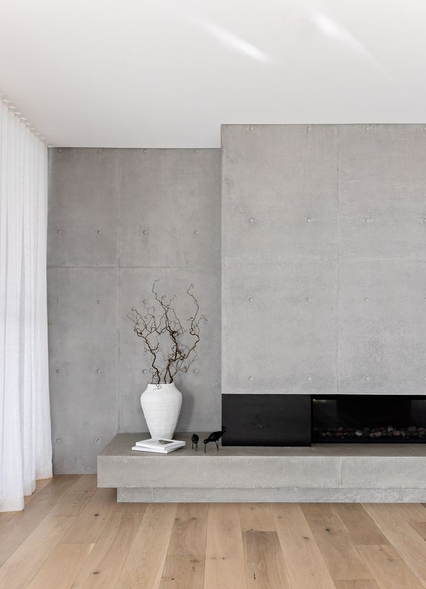 Minimalist living room featuring a full-height exposed concrete feature wall with visible form-tie marks, a black built-in linear fireplace, and a projecting concrete hearth. Pale timber plank flooring, sheer white curtains, and a sculptural white vase create a restrained, monochromatic interior.