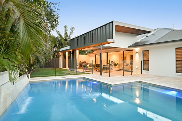 Exterior architectural photograph of a contemporary tropical residence at dusk, viewed across a large swimming pool. The home combines a white rendered volume with a dark vertical-clad projecting upper form, broad open glazing, slender dark posts, warm timber soffits, and a pitched metal roof. Palm vegetation surrounds the landscaped pool area, while the illuminated open-plan interior is visible through the glazing.