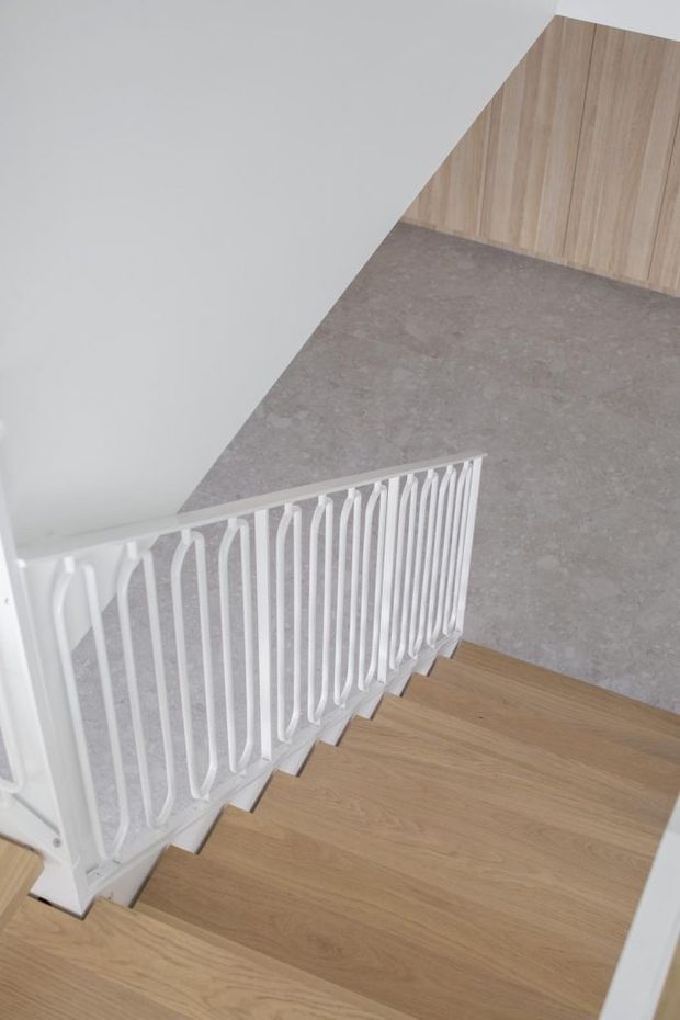 A bright minimalist stairway viewed from above, with light timber stair treads, a distinctive white sculptural metal balustrade, pale grey flooring, white walls, and warm timber wall panelling at the lower level.