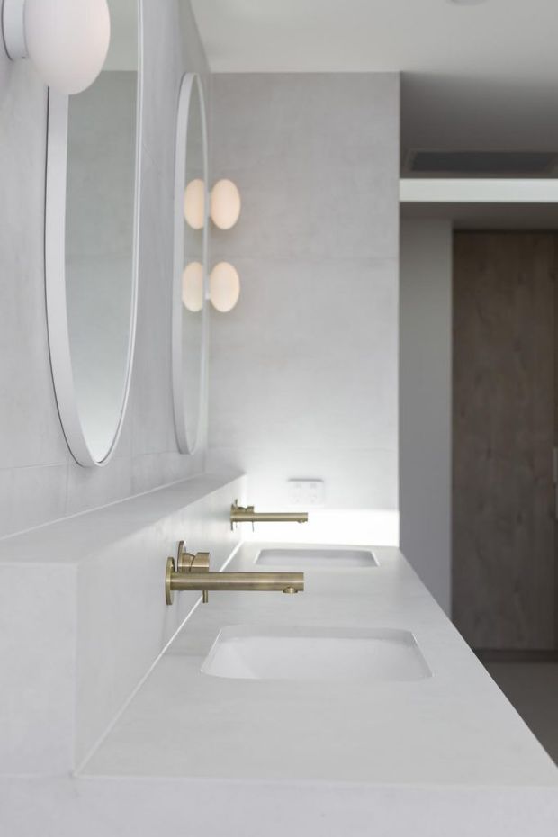 A bright, minimalist bathroom vanity with a long white benchtop, two integrated rectangular basins, wall-mounted brushed brass tapware, large rounded-oval mirrors, and soft globe wall lights. White and pale grey surfaces create a clean contemporary interior, with a dark timber door visible beyond.