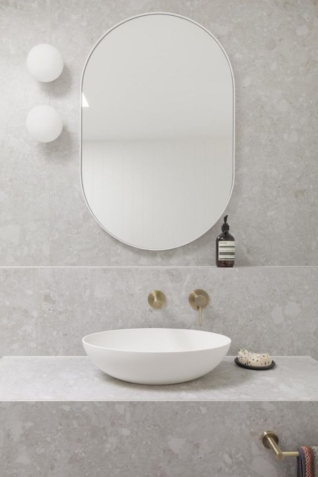 A minimalist bathroom vanity vignette featuring a large rounded-arch frameless mirror, two spherical wall lights, a white vessel basin on a pale grey stone vanity, and brushed brass wall-mounted tapware and accessories. The walls and benchtop are clad in matching light grey stone or porcelain slabs, creating a calm, refined contemporary interior.