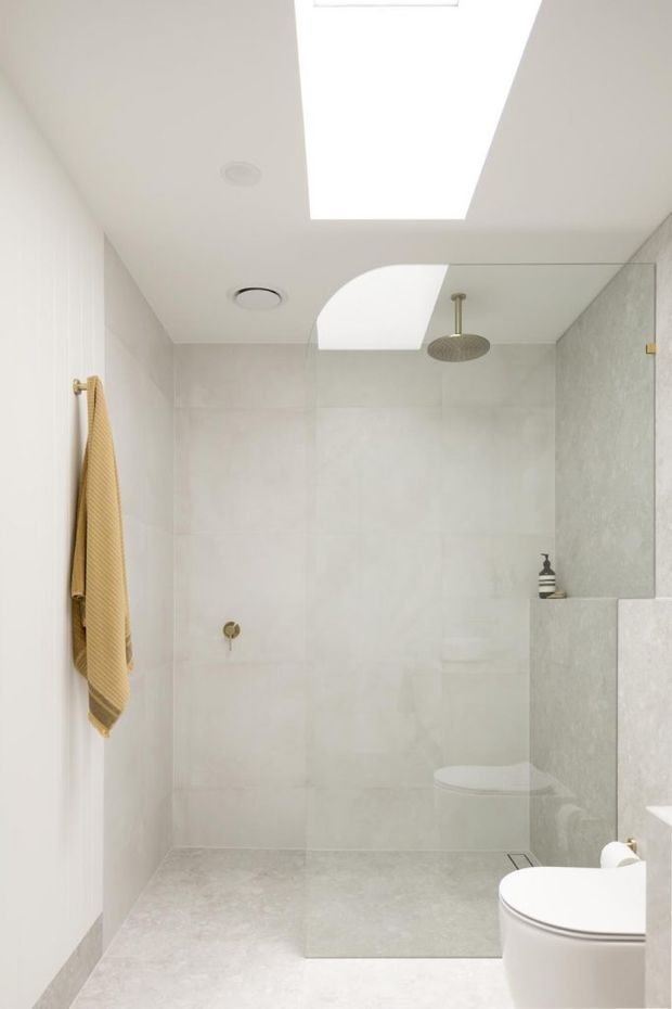 Bright, minimalist contemporary bathroom with a frameless glass walk-in shower, large pale grey stone-look wall and floor tiles, a wall-mounted rainfall shower, recessed ceiling ventilation, and a skylight. A white toilet is visible in the foreground, with a warm mustard towel adding a small accent.