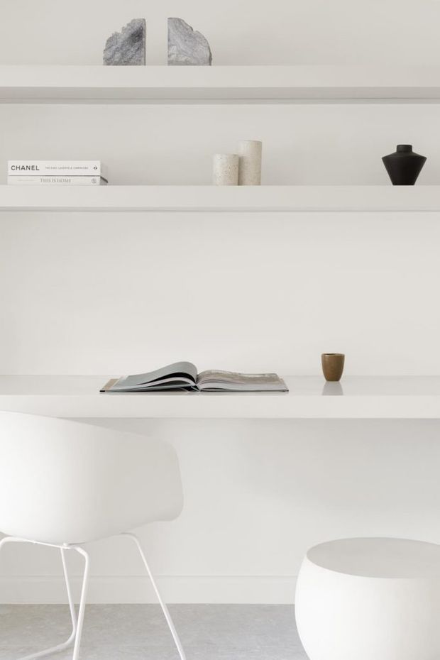 Minimalist white built-in shelving and desk-like joinery in a bright interior, styled with a few books, sculptural stone pieces, ceramic vessels, and an open magazine. A white chair and rounded white side table sit against the shelving, with a pale grey floor completing the calm monochromatic scheme.